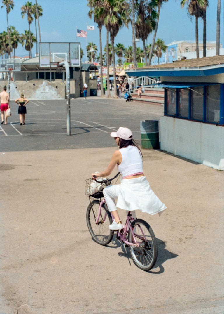 A woman rides a bicycle along a palm tree-lined path near a beachside basketball court.