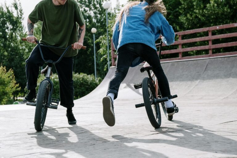 Two BMX riders performing stunts on their bikes at an outdoor skatepark on a sunny day.